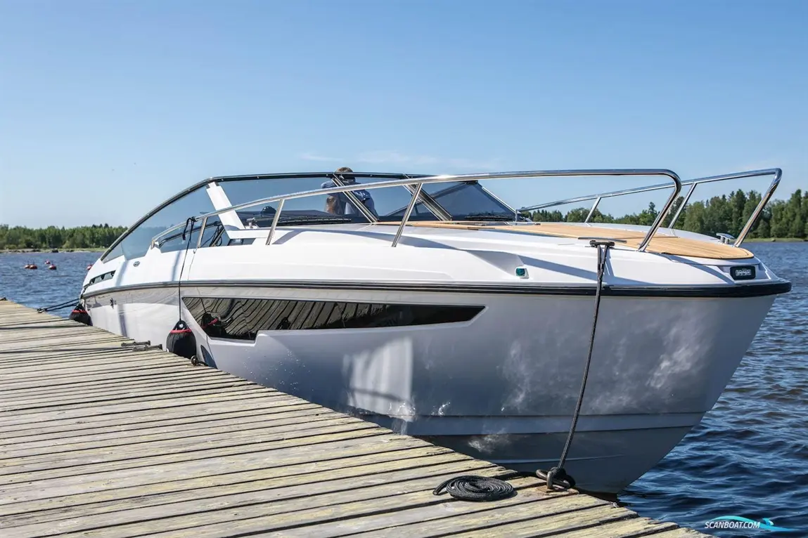 A sleek white boat is docked alongside a wooden pier, with blue water and trees in the background on a sunny day.